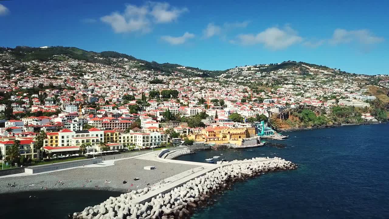 Aerial view highlighting the vibrant cityscape of Funchal, Madeira, with a bustling harbor, colorful buildings, and majestic mountains under a sunny, partly cloudy sky, drone establishing shot