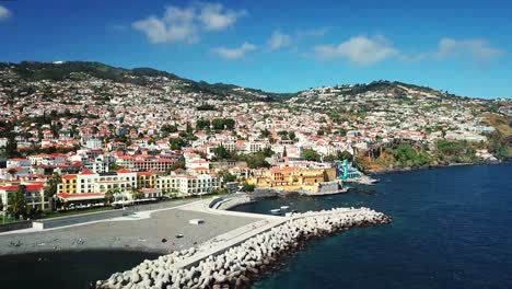 aerial view highlighting the vibrant cityscape of funchal, madeira, with a bustling harbor, colorful buildings, and majestic mountains under a sunny, partly cloudy sky, drone establishing shot