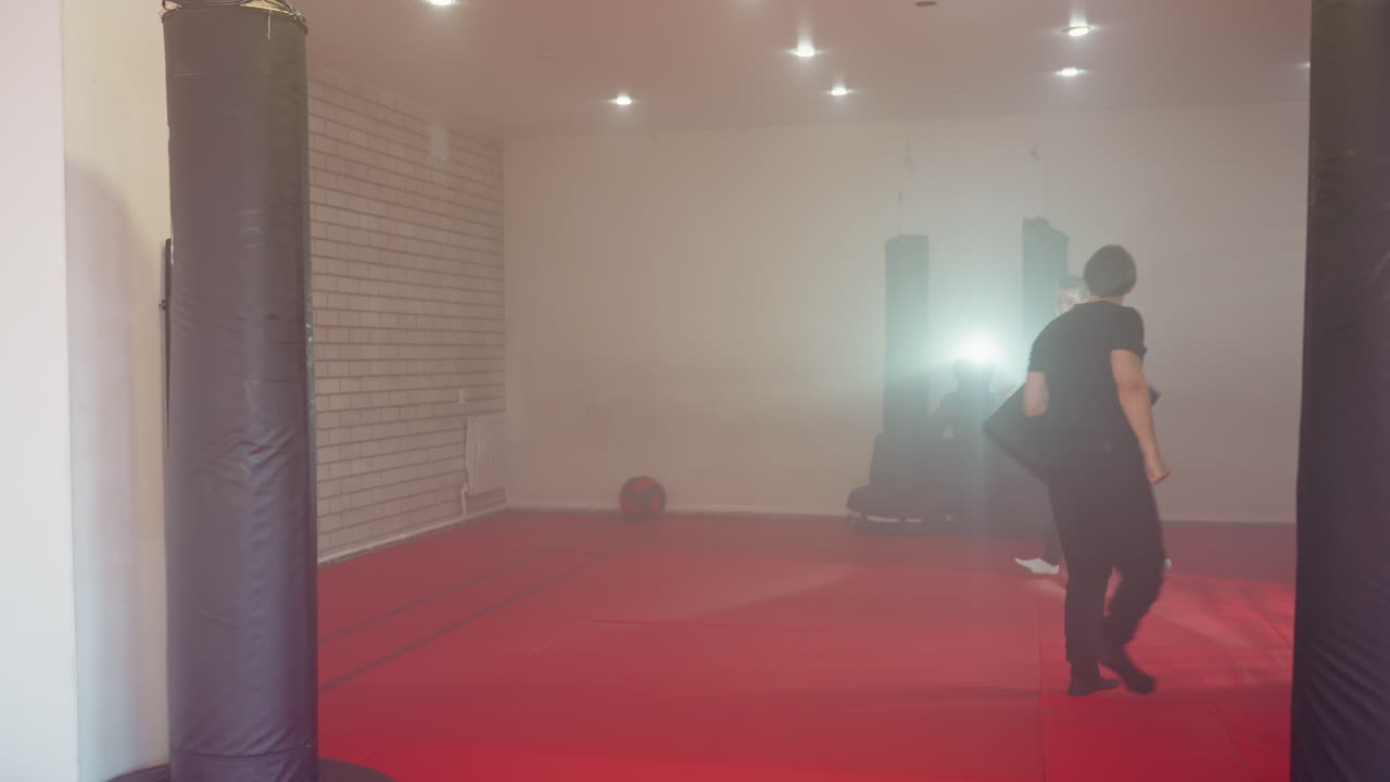 Two men training martial arts in gym with punching bags, one holding shield for protection while other prepares to strike, red mats on floor, foggy atmosphere, intense practice, spotlight glare in background