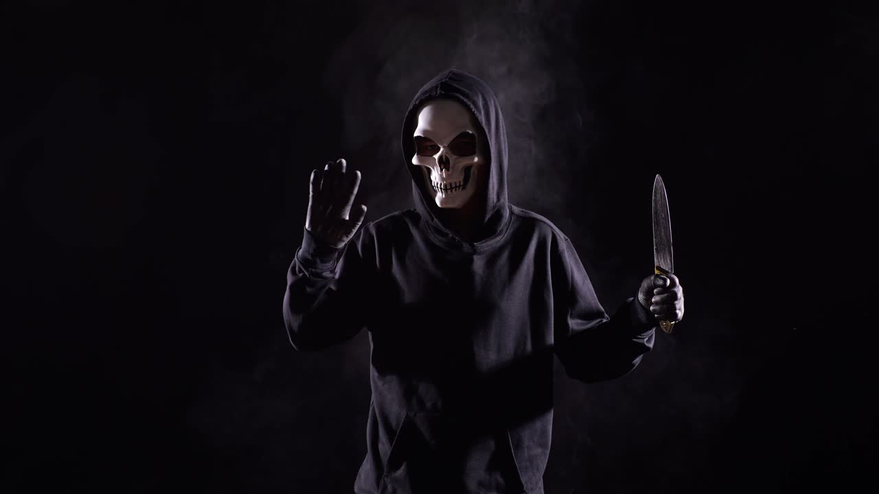 Scary Man In The Hooded Sweatshirt Wearing Halloween Mask Making Bye Gesture While Holding A Knife On The Black Background With Smoke