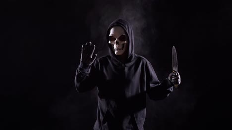 scary man in the hooded sweatshirt wearing halloween mask making bye gesture while holding a knife on the black background with smoke