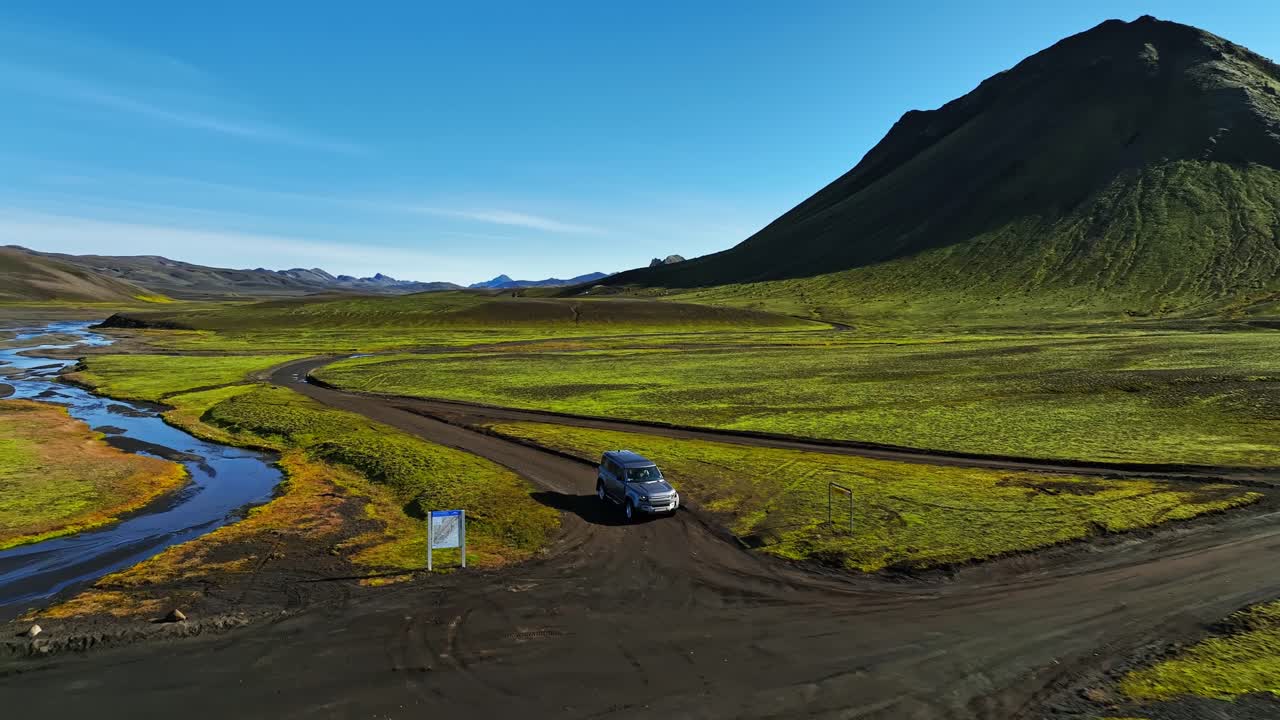 Veduta aerea con drone di un SUV che percorre una strada sterrata tortuosa accanto a un fiume negli altopiani verdi dell'Islanda, con alle spalle una drammatica montagna vulcanica sotto un cielo azzurro.