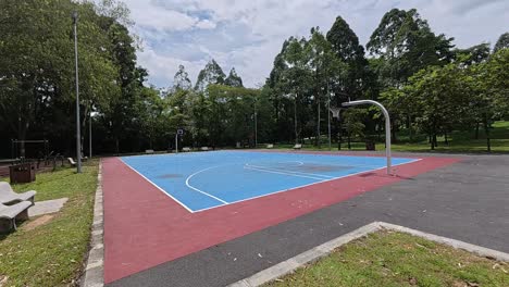 an empty basketball court in a tropical park, surrounded by lush green trees under a bright, sunny sky. perfect weather.