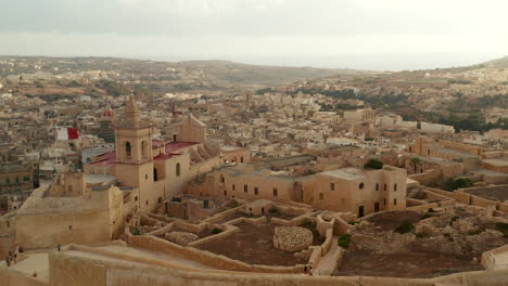 Fuerte-Del-Castillo-De-Gozo-Con-Castillo-Ondeando-La-Bandera-De-Malta-En-Un-Hermoso-Color-Marrón-Arena,-Tobogán-Aéreo-A-La-Derecha