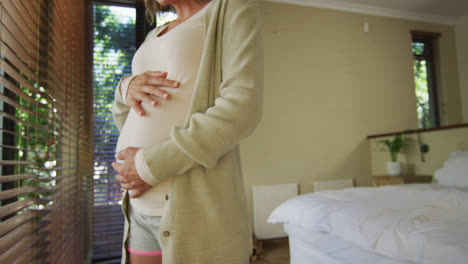 midsection of caucasian pregnant woman standing at window and touching belly