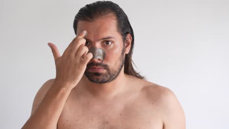 joven metrosexual con barba sonriendo aplicando una máscara facial de arcilla carbonífera mirando en el espejo del baño