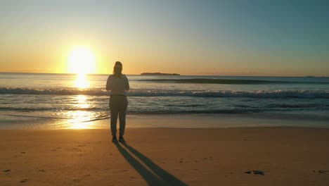 Woman-stands-alone-at-the-beach-looking-at-the-ocean-waves-and-sunrise-at-golden-hour