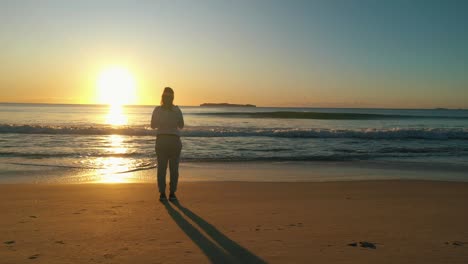 woman stands alone at the beach looking at the ocean waves and sunrise at golden hour