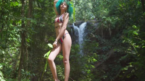 a young woman stands at the base of a waterfall in a bikini and green hair