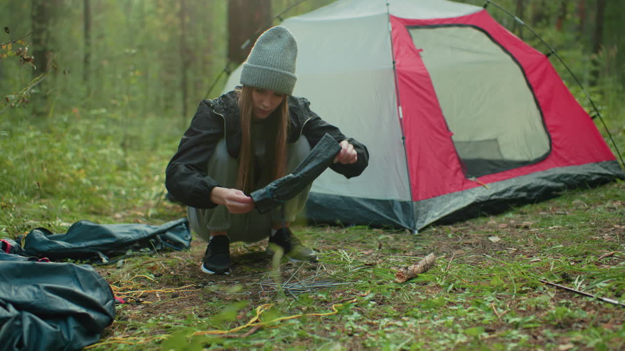 young camper in gray beanie crouches to pick up scattered camp equipment dropped on forest floor near pitched tent during peaceful outdoor morning surrounded by trees and soft light