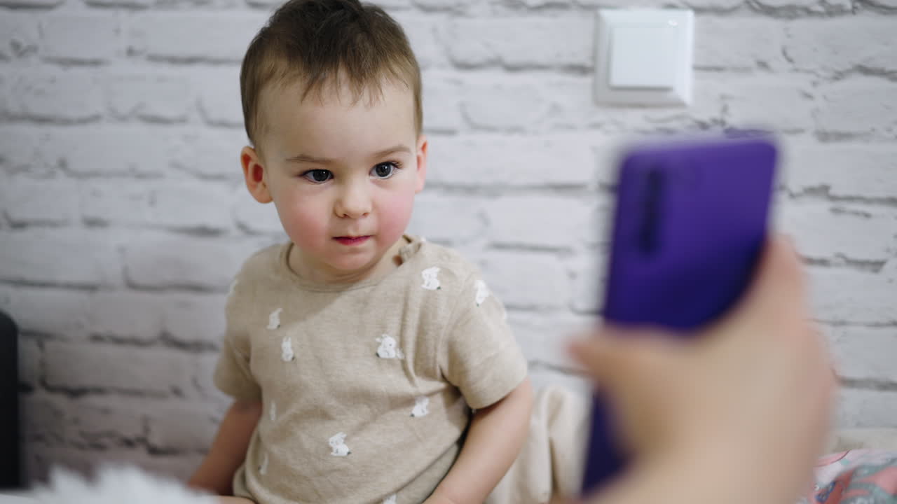 Beautiful toddler boy with cute rosy cheeks is focused on the phone. Parent holds a gadget for kid and he is mesmerized by the view.