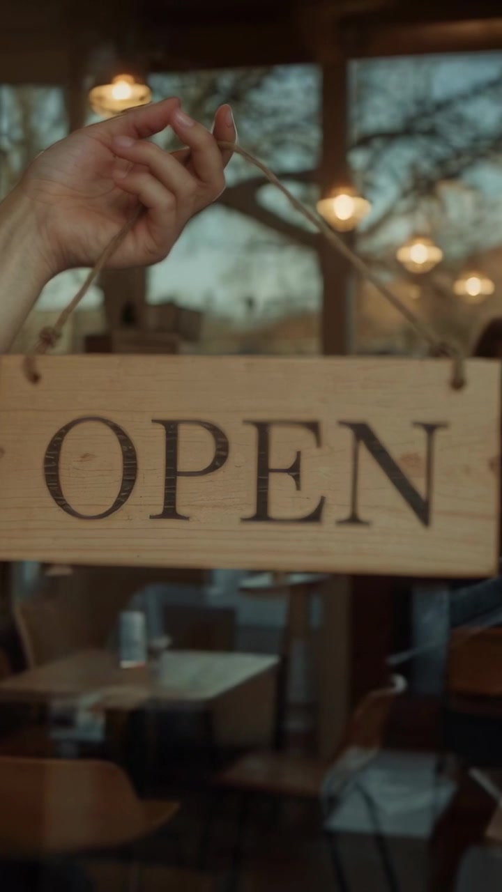Video verticale: Una mano che entra e afferra una corda posiziona un cartello "OPEN" sulla vetrina di un caffè, mostrando lo stato di apertura.