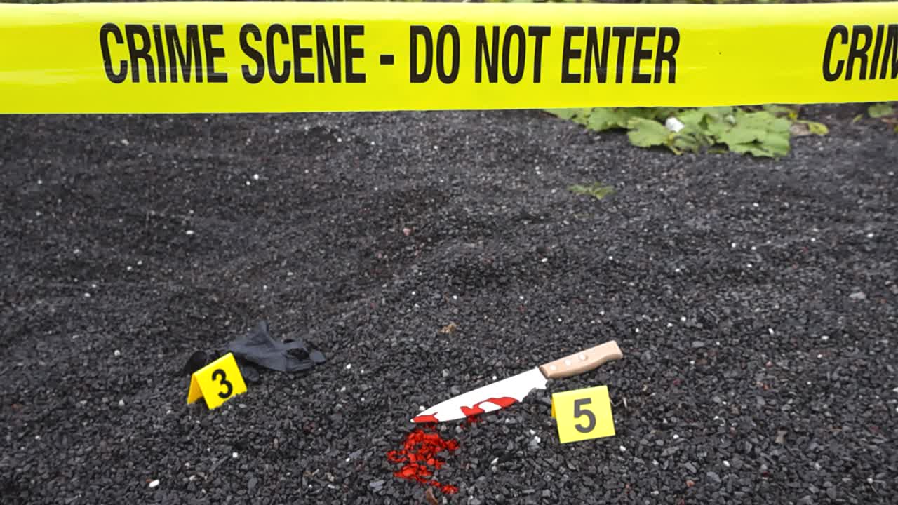Yellow police line or forensic investigation crime scene tape in front of police markers that are next to evidence like bloody murder weapon knife and nylon black glove on dark colored gravel ground
