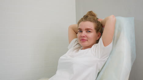 woman with tied hair sitting on salon chair with hands supporting head, leaning back near gray and white wall. she appears relaxed and calm in well-lit modern setting with minimal decoration
