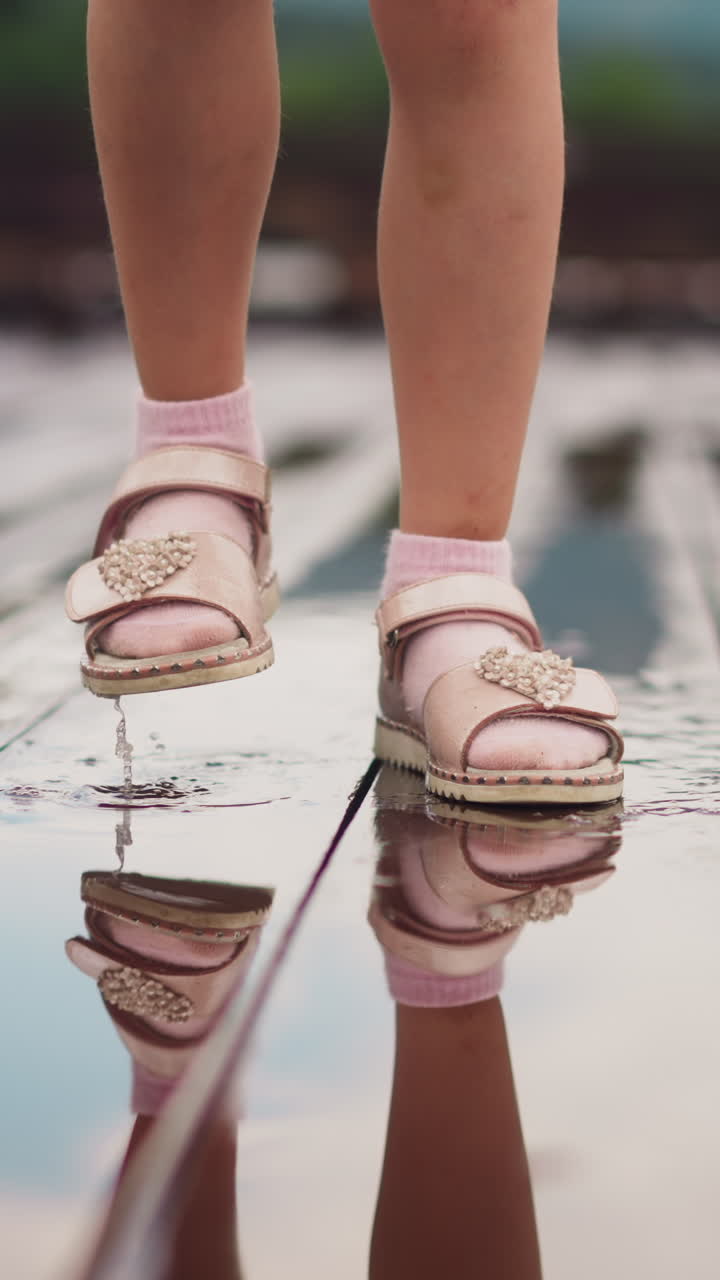 Little girl in sandal shoes and socks stamps feet in small puddle under drizzling rain playing on empty wooden deck close view slow motion