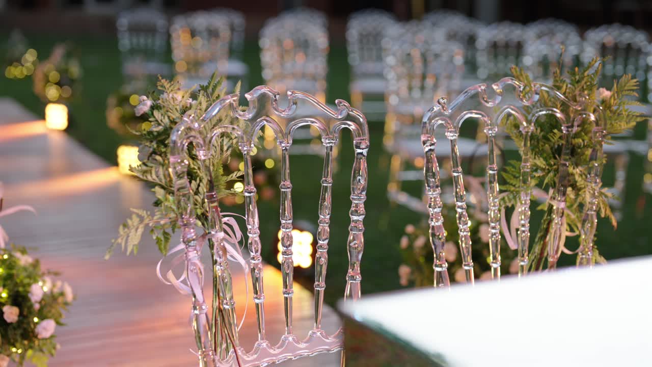Primo piano dell'allestimento per una cerimonia di matrimonio all'aperto con navata decorata, composizioni floreali ed eleganti sedie trasparenti.