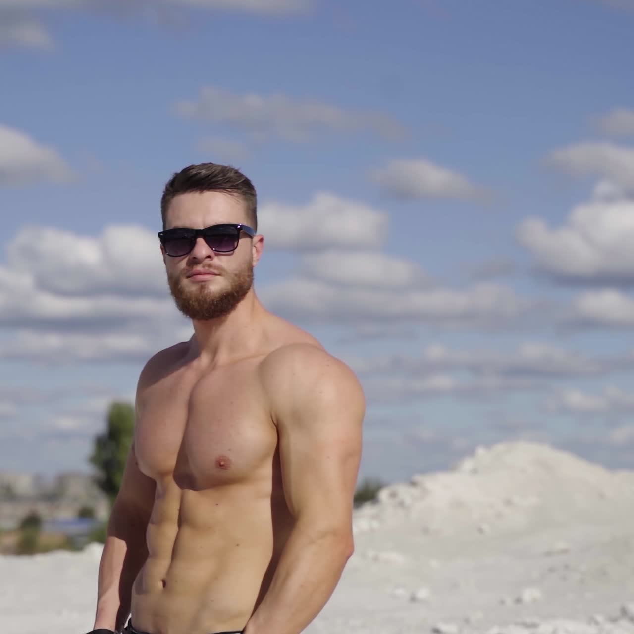 Portrait of athlete at sunlight. Handsome shirtless man in black sunglasses standing on the top of white hill against the beautiful summer sky. Healthy lifestyle.