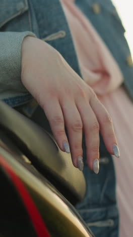 primer plano de la mano de una mujer con uñas pintadas descansando en un asiento de motocicleta, ella lleva una chaqueta de vaqueros y un vestido rosa, con un fondo suavemente borroso