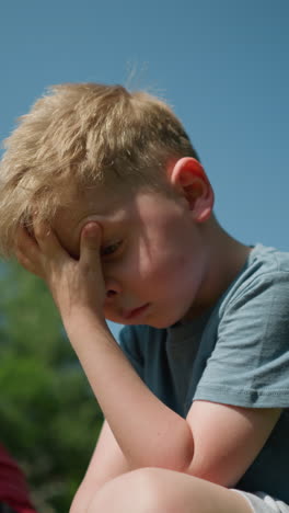 a tired-looking little boy wearing a blue shirt rubs his face with his left hand, with a blurred view of his elder brother looking at him
