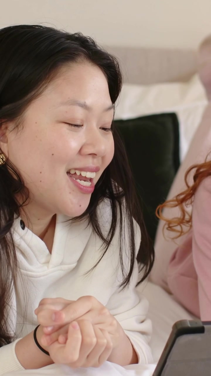 Video verticale: Donna asiatica sorridente in felpa bianca con cappuccio, con le mani giunte, che reagisce a un tablet sul letto.