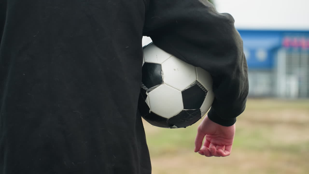 Back view of someone in a black outfit holding a classic black and white soccer ball under their arm, with a blurred grassy field and a blue building in the background