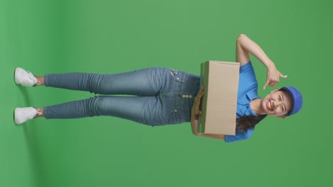 full body of asian female courier in blue uniform showing call me gesture and smiling while delivering a carton on green screen background in the studio