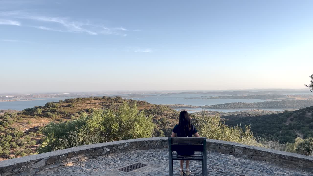 woman sits, admiring the Alentejo landscape at sunset with a river winding through hills and mountains, under a sky of warm orange and purple hues.