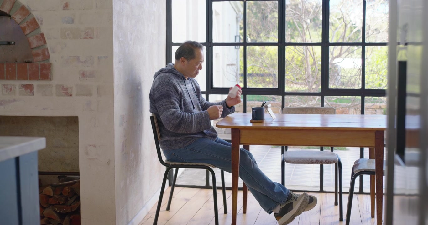 Asian man sitting at table by window, ending call, opening meds bottle while checking tablet