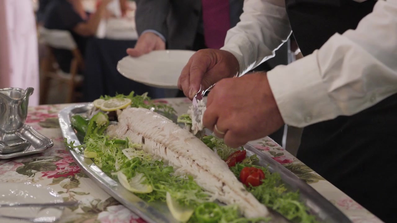 Un cameriere che indossa una fede nuziale porziona un pesce intero al forno con insalata, limone e pomodori su un vassoio d'argento, tenendo un piatto per un ospite