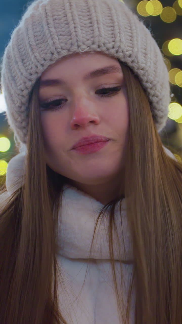 Close-up of woman in cozy winter coat and beanie walking toward a seat with head slightly bowed, festive city lights glow in background as people skate and socialize