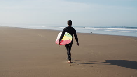 campo lungo di un surfista maschio con la gamba artificiale che tiene la tavola da surf sotto il braccio e cammina lungo la spiaggia