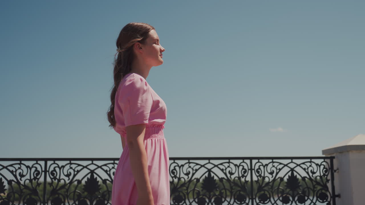 Mulher jovem de vestido rosa em pé perto de um corrimão de ferro ornamentado em um calçadão banhado pelo sol, cabelo trançado esvoaçando na brisa, fotos de perfil cinematográficas capturam o humor contemplativo e cenas estéticas vintage