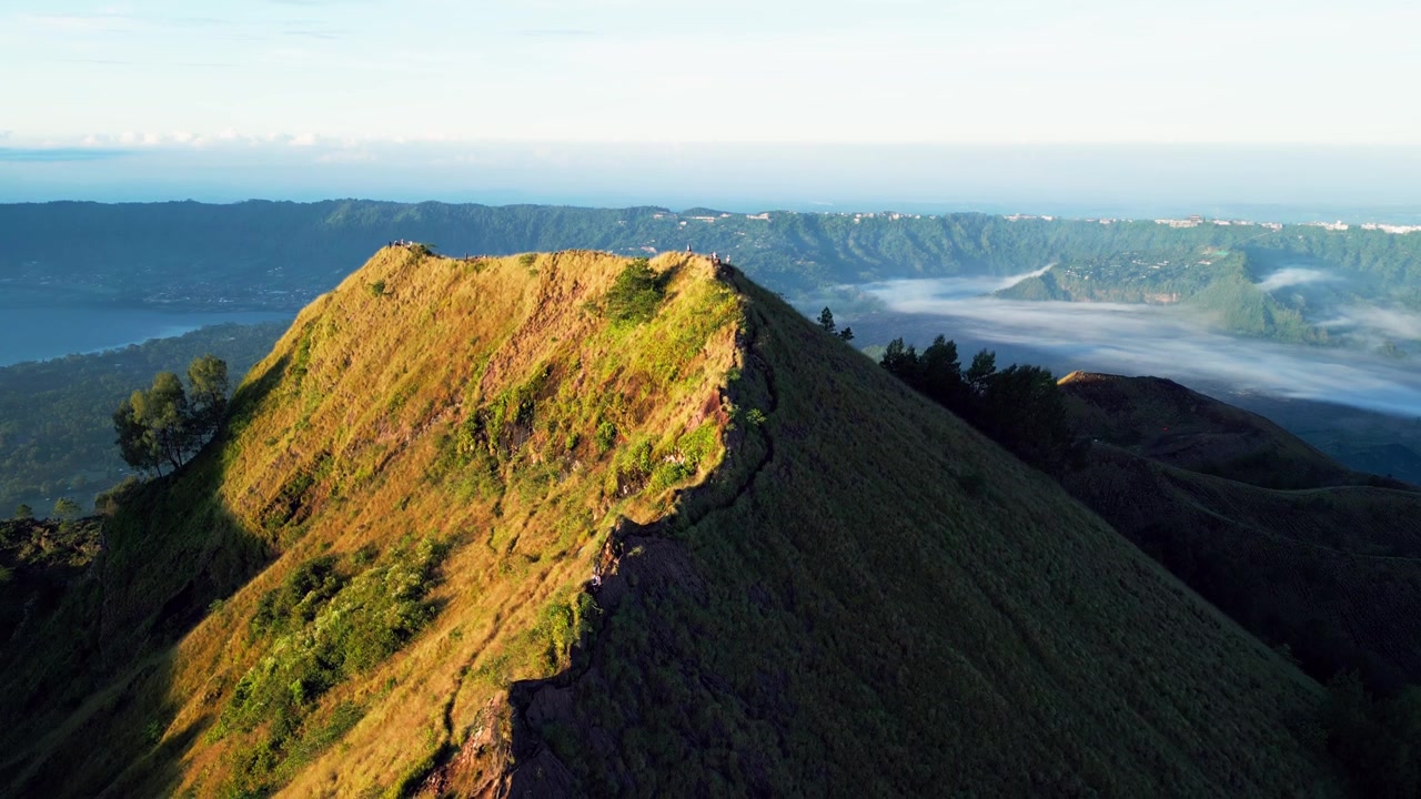 Trekking all'alba con escursionisti che attraversano una stretta cresta vulcanica che si affaccia su una valle caldera coperta di nebbia e sugli altipiani del Monte Batur nella calda luce del mattino, ripresa con drone in discesa.