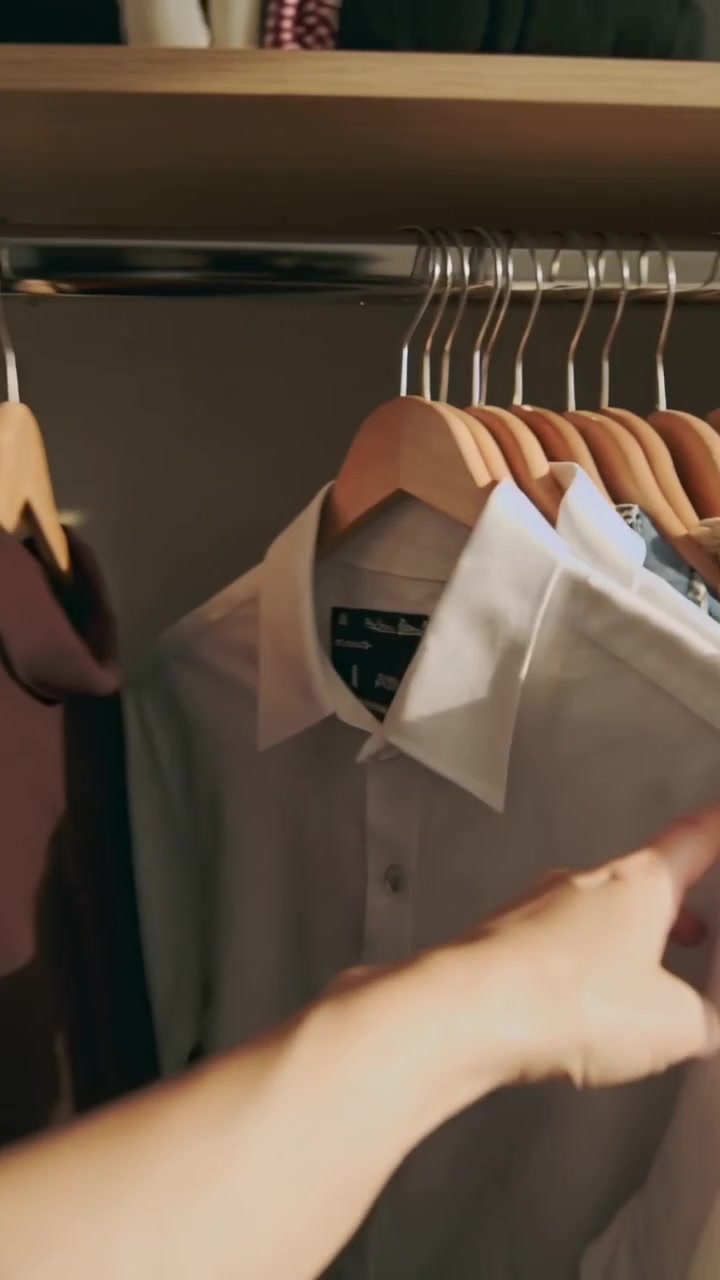 Vídeo vertical: Mãos deslizando com manga neutra, organizando camisas em armário de quarto, cabides de madeira.