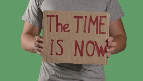 man holds in front of him a poster from a cardboard box with the words the time is now. protest against police brutality terror and racism. isolated a green screen, chroma key. close up. slow motion