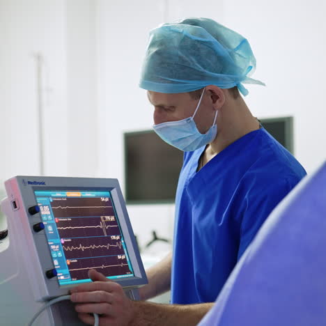 uniformed male medic in mask and cap standing near the monitor of lung ventilator. surgical operation in hospital.