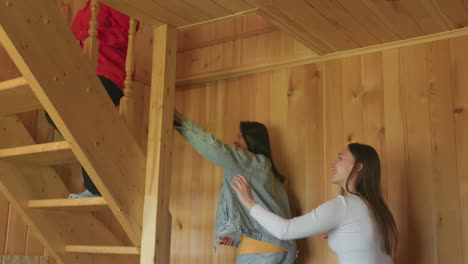 three young women inside cozy wooden interior as girl in red leads others toward staircase, casual indoor moment showing friendship, navigation, movement, and curiosity in warm natural cabin setting
