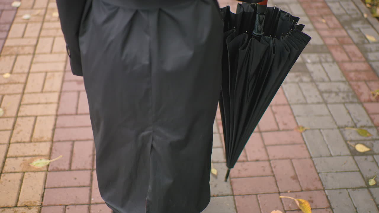 Rear view of person in black coat holding closed black umbrella with wooden handle while walking on wet paved sidewalk with fallen leaves, symbolizing anticipation, preparation, weather protection