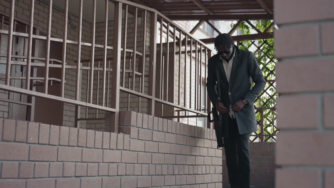 Hombre de piel oscura con gafas de sol abrochándose el abrigo mientras baja una escalera exterior junto a paredes de ladrillo, barandillas de hierro y vegetación enrejada, mostrando un ambiente informal de negocios y un humor de camino al trabajo matutino