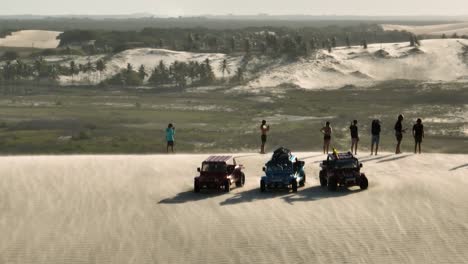 aerial orbiting view of a group standing on the edge of a sand dune near off road vehicles overlooking the dramatic views as strong wind blows sand across the face of the dune