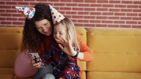 Madre-Sentada-Con-Su-Hija-Preescolar-Con-Gorras-De-Cumpleaños,-Tiene-Videollamada-Por-Móvil