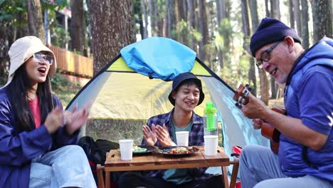 happy asian father, son and daughter playing guitar and singing together at campsite