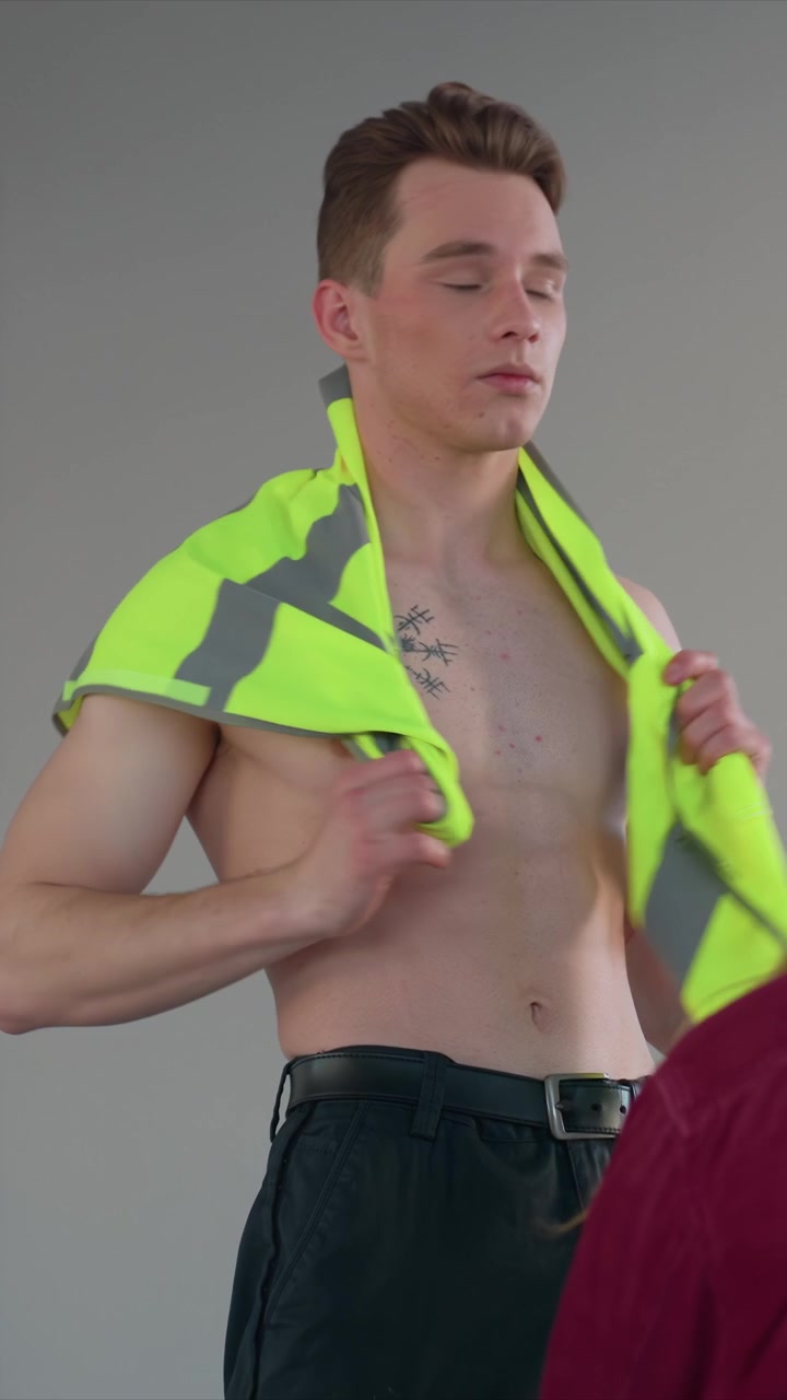 Uomo tatuato in un ambiente grigio semplice tiene in mano un neon. Il tatuaggio presenta forme angolari centrate intorno alla zona del collo. Scena interna con sfondo grigio semplice e illuminazione uniforme.