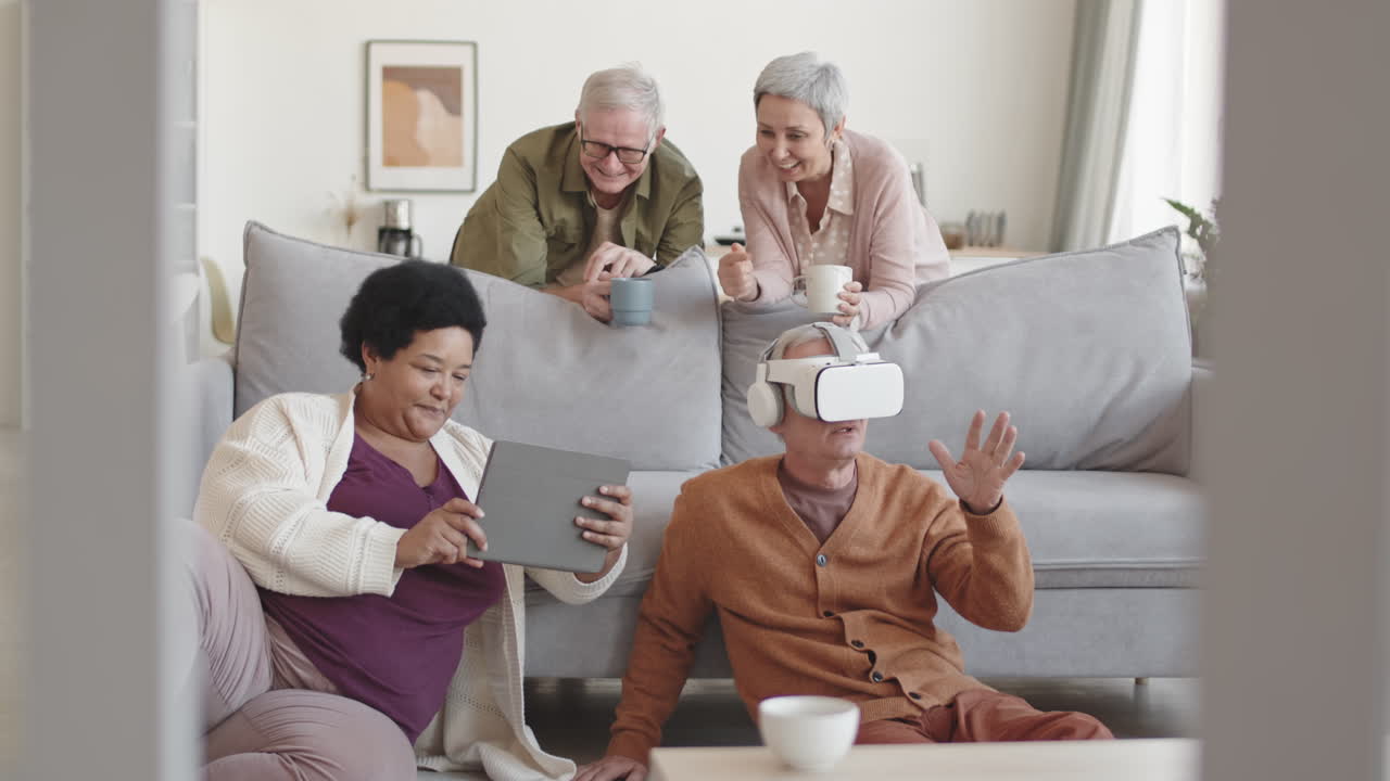 Medium long of happy diverse senior friends hanging out around couch, man using VR headset, African woman having tablet computer, grey-haired couple holding cups looking at them