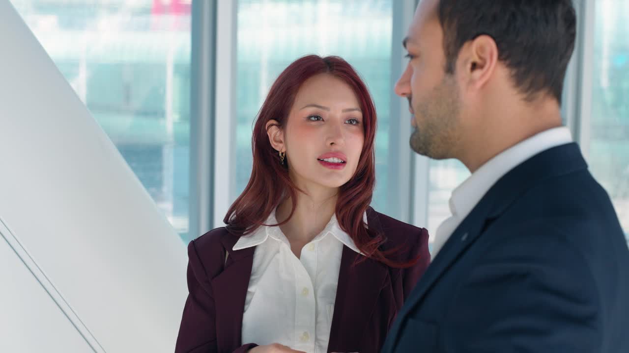 明るいモダンな企業回廊で、バーガンディ色のスーツを着た若い女性ビジネスパーソンが、クローズアップのスローモーションショットで男性同僚に自信を持って話す