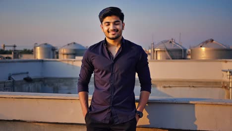 confident young engineer standing on rooftop with hands in pockets, industrial silos visible in the background during sunset, showcasing professionalism and industrial environment
