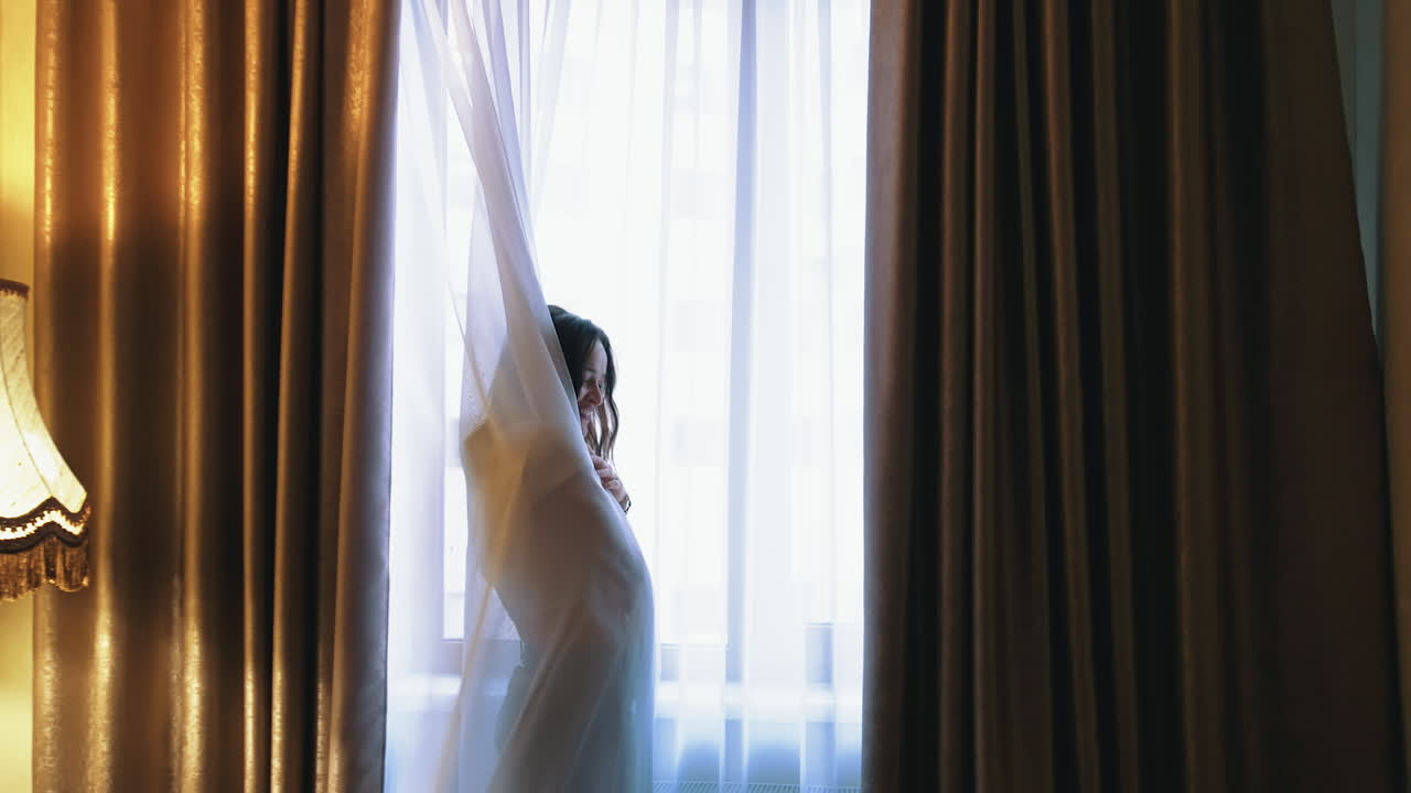 Happy woman enjoys in a comfortable hotel. Portrait of a cheerful girl near the large window curtains. Smiling woman feeling joy standing in luxury apartment.
