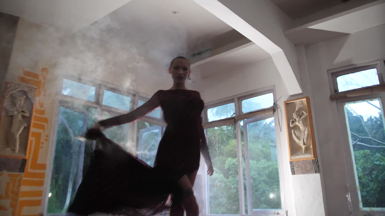 A mysterious young woman with shaved head is holding a smoke grenade and dancing dramatically back and forth barefoot while spreading the smoke around the room
