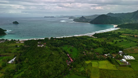 An aerial shot reveals picturesque Selong Belanak Beach, with its sandy coastline, turquoise waters, and lush green mountain hills in the distance