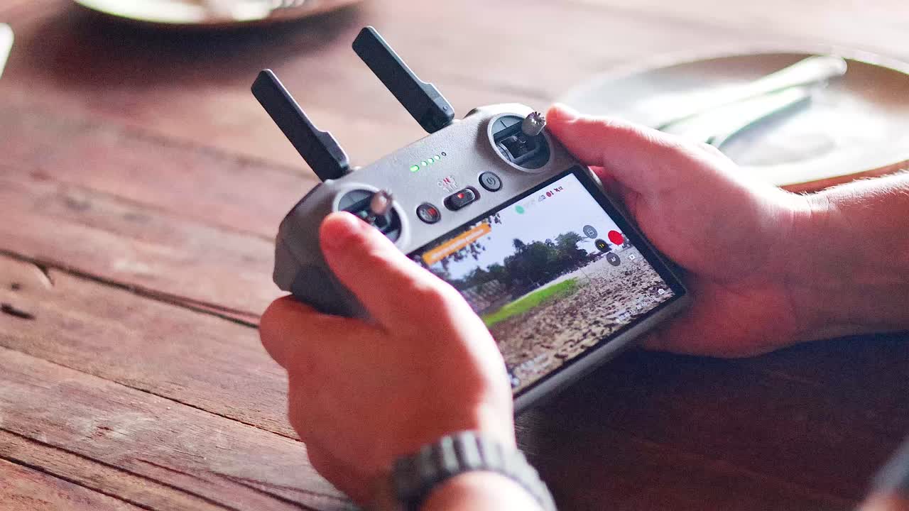 Hands operating a drone controller indoors with a screen displaying aerial views. Natural lighting, wooden table setting